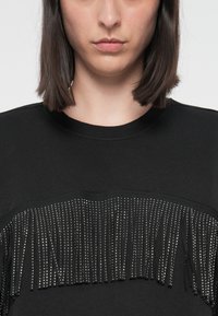 Zwarte sweatshirt met een franjes ontwerp en decoratieve zilveren studs langs het borstgebied, geribbelde halslijn en een zachte stofstructuur.
