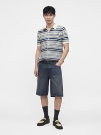 Jeune homme debout portant une chemise à rayures à manches courtes boutonnée, un short long en denim, des chaussettes noires et des mocassins bicolores.