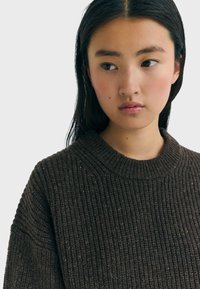 Giovane donna con lunghi capelli scuri che indossa un maglione in maglia marrone scuro, che guarda leggermente di lato su uno sfondo chiaro e semplice.