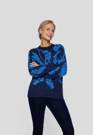 Blue patterned sweater with a navy base, featuring a soft texture and loose fit. Model is wearing navy pants, arms crossed, smiling.
