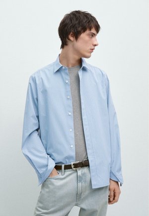 Junger Mann, der ein hellblau gestreiftes Hemd über einem grauen T-Shirt, hellblaue Jeans und einen braunen Gürtel trägt, nach rechts blickend, mit einer Hand in der Tasche.