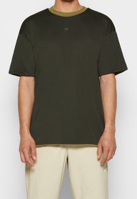 Camiseta de manga corta verde oscuro con una tela texturizada, contrastada por un cuello de oliva claro. Presenta un pequeño logo negro en el pecho.