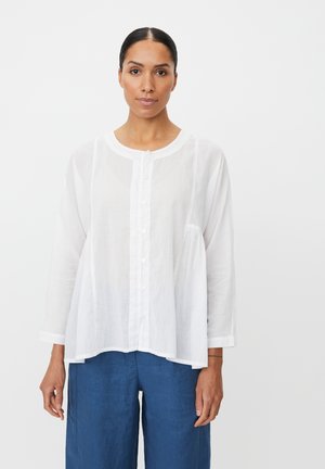 Hvid, langærmet bluse lavet af letvægtsstof med rund halsudskæring og forsideknapper. Kombineret med blå linnedbukser.