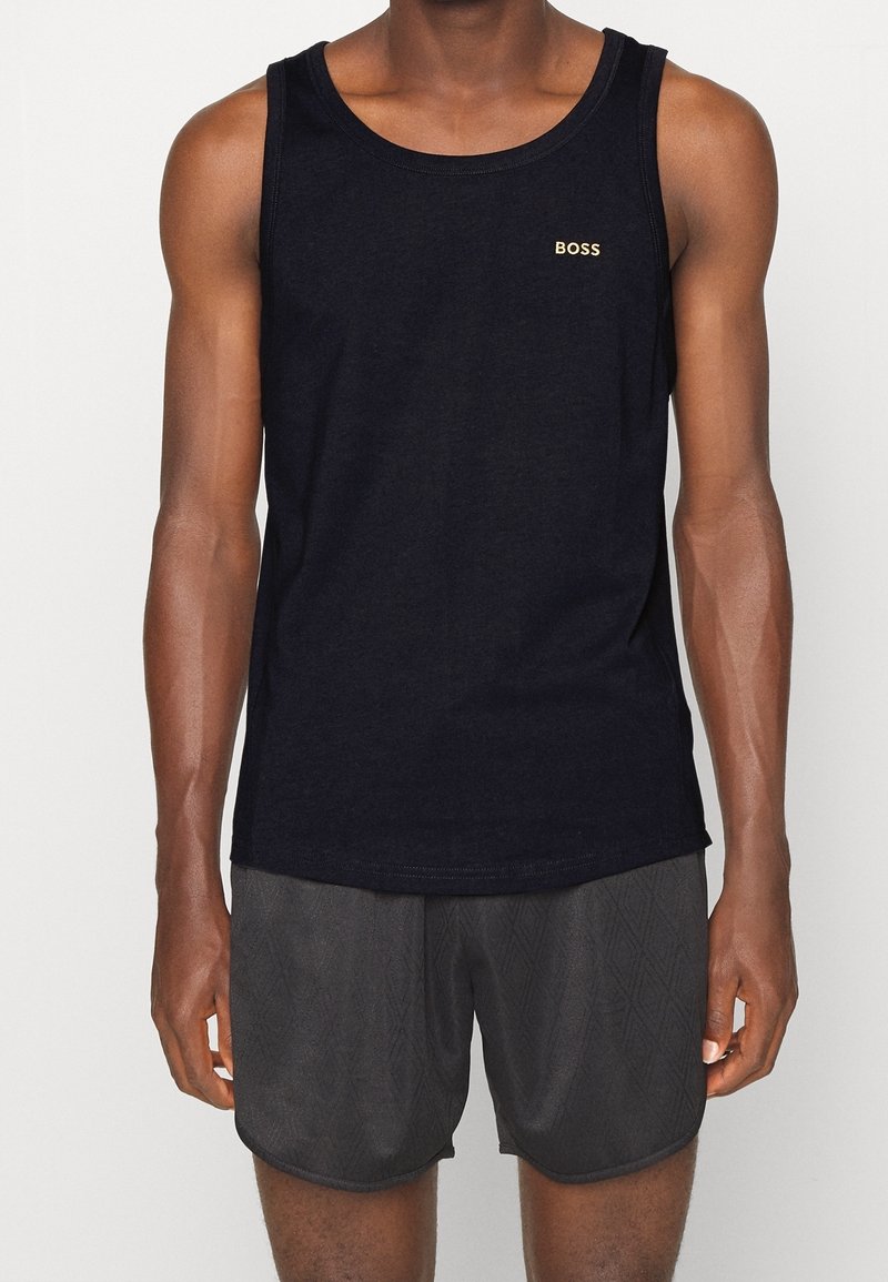 Homme portant un débardeur noir sans manches avec le logo « BOSS » et un short de sport texturé foncé, debout devant un fond uni.