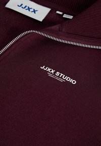 Бордо суитшърт с цип и рипсено яке. Има бял лого „JJXX STUDIO“ и гладка текстура. Контрастен сребърен детайл на ципа.