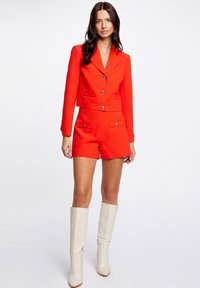 Blazer rosso cropped con due tasche frontali e pantaloni corti rossi abbinati. Modella che indossa stivali al ginocchio color crema. Tessuto liscio, vestibilità strutturata.