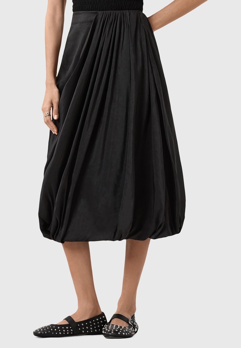 Jupe boule noire à plis portée par une personne debout, associée à des ballerines noires cloutées et un haut noir.