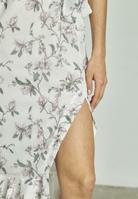 Das Blumendruckkleid besteht aus einem weißen Stoff mit hellrosa und grünen Blumen. Der Schlitz an der Seite und der ausgestellte Saum verleihen dem Kleid Details und Textur.