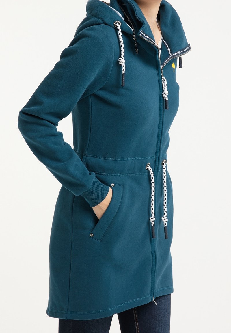 Hoodie zip-up in teal realizzato in tessuto morbido, con cappuccio regolabile, tasche laterali e polsini elastici con dettagli neri e hardware in metallo.