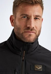 Black jacket featuring a high collar, zip closure, and logo patch. Smooth texture with stitching details and gold accents on the zipper.