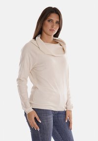 Maglione color crema con collo a cowl, maniche lunghe e una struttura morbida, abbinato a jeans in denim aderenti. Design semplice e moderno.