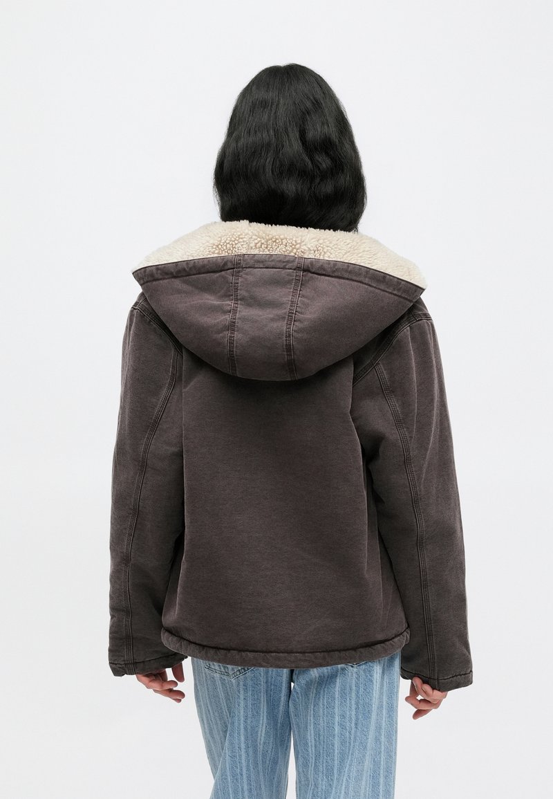 Brown hooded jacket with a shearling collar, relaxed fit, and visible stitching details, worn over light blue striped jeans.