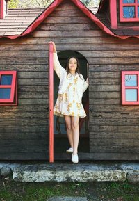 Casa de madera con ribetes rojos, que cuenta con una puerta redondeada y ventanas coloridas. Una niña con un vestido floral se encuentra en la entrada.