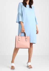 Růžová kožená taška přes rameno s dvojitými uchy a zlatými akcenty, doplněná světle modrými šaty s rozšířenými rukávy a bílými sandály na podpatku.