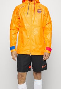 Ljust orange zip-jacka med FC Barcelona-logotyp, blå och röda detaljer på ärmarna och präglat textmönster. Kombinerad med svarta shorts och vita strumpor.
