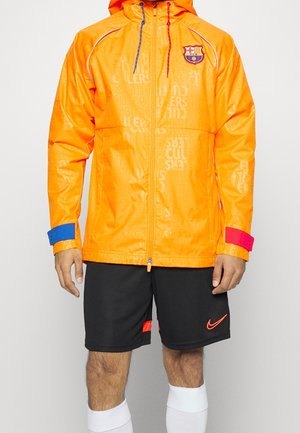 Giacca zip-up arancione brillante con logo del FC Barcelona, dettagli blu e rossi sui polsini e motivo di testo in rilievo. Abbinata a pantaloni corti neri e calze bianche.