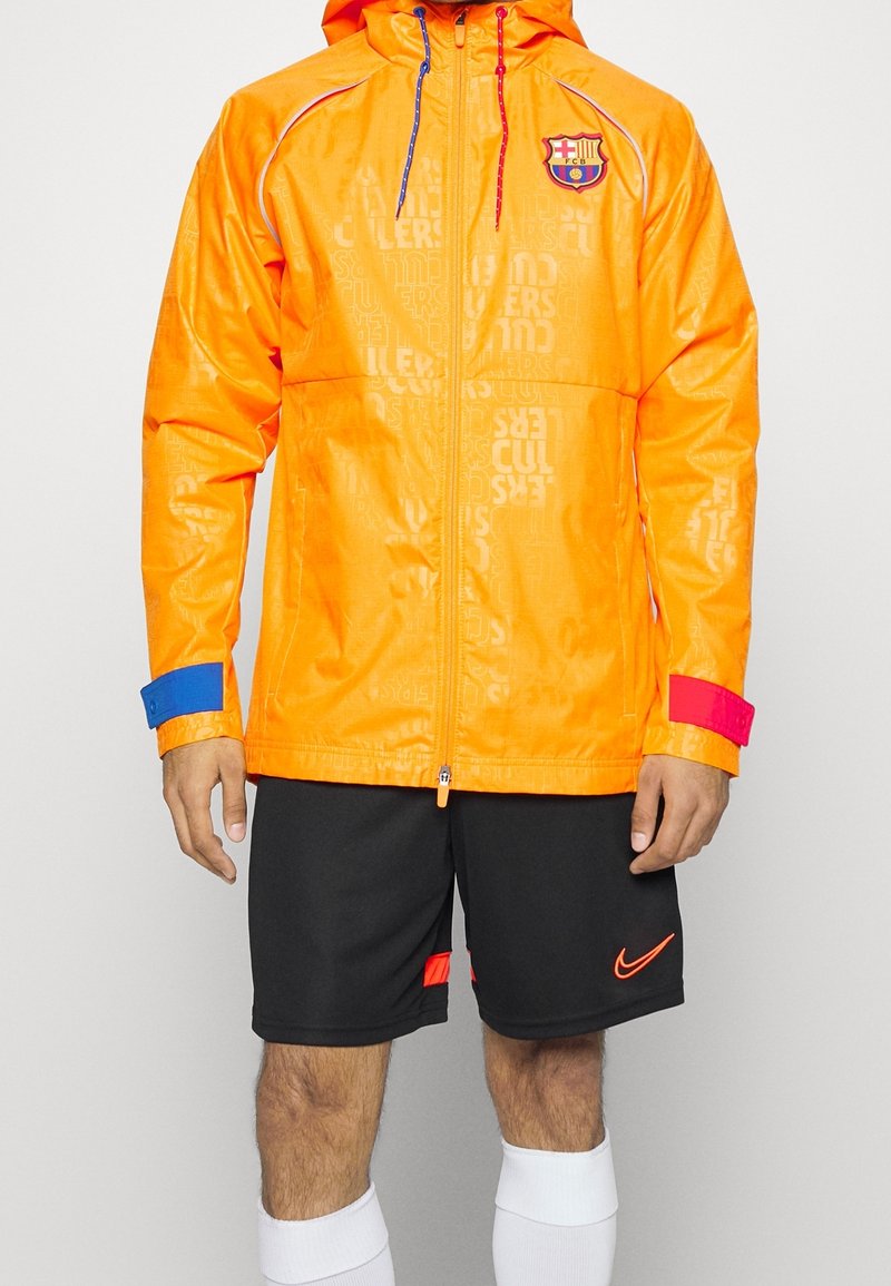 Ljust orange zip-jacka med FC Barcelona-logotyp, blå och röda detaljer på ärmarna och präglat textmönster. Kombinerad med svarta shorts och vita strumpor.