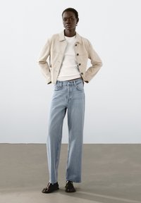 Beige mockajacka med knäppning fram, vit t-shirt under, ljusblå raka jeans och svarta sandaler. Slät textur genom hela plagget.