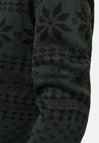 Knit sweater in dark green with a black floral pattern. Features a textured finish and ribbed cuffs. Close-up of the sleeve detail.
