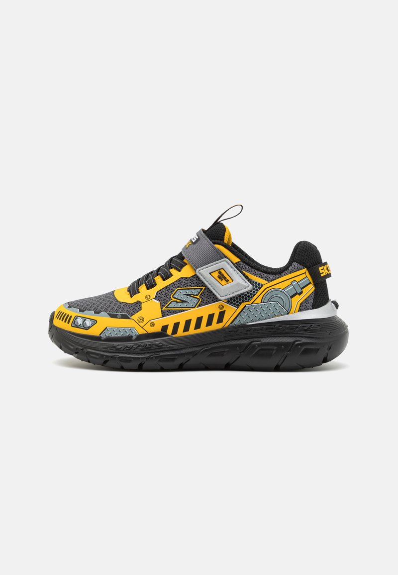 Svart og gul sneakers med mesh-overdel, mønstrede detaljer, forsterket tå og gummisåle. Har en spenn og teksturerte detaljer.