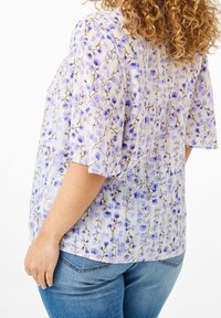 Haut léger à motifs floraux avec des fleurs violettes sur fond blanc, manches courtes et coupe ample. Porté avec un jean bleu.