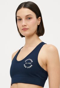 Marineblå sports-bh laget av stretchy materiale, med et sirkulært hvitt logo som har teksten "HILFIGER" og "NEW YORK", samt en racerback design.