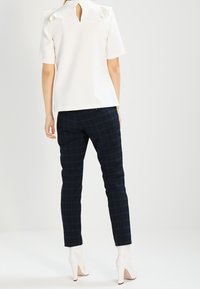 Vit kortärmad topp med en nyckelhålsöppning bak, kombinerad med mörkblå rutiga byxor och vita högklackade skor. Slät textur överlag.