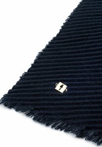 Ciemnoniebieska materiał z żebrowanym wzorem, z frędzlami na brzegach i małym kwadratowym złotym emblematem z abstrakcyjnym symbolem w pobliżu jednego z rogów.
