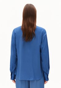 Persoon met lang bruin haar draagt een losse blauwe shirt en bijpassende broek, getoond vanuit de rug tegen een effen witte achtergrond.