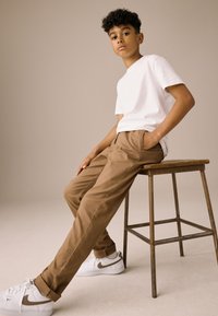 Tricou alb cu mâneci scurte, pantaloni bej cu croială lejeră și adidași albi cu accente bej. Stă pe un scaun din lemn, având un fundal neutră.