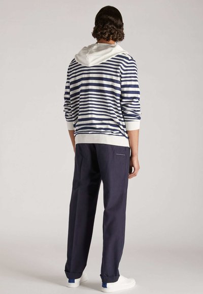Gestreifter navy- und weißer Hoodie mit einer weißen Kapuze, kombiniert mit navyfarbigen Hosen. Die Hosen haben einen dezenten Akzent auf der Gesäßtasche und umgeschlagenen Säumen.