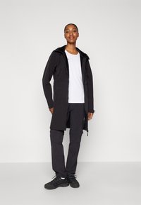 Veste noire longue avec capuche et fermeture éclair, associée à un pantalon sombre et des chaussures noires. La veste possède des cordons de serrage et une texture lisse.