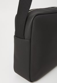 Sort rektangulær taske med tekstureret overflade, justerbar rem og kontrasterende lynlåsdetalje. Glat finish med en lille sidelomme.