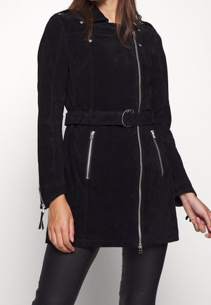 Kvinde iført en sort ruskindjakke med bælte, asymmetrisk frontlynlås og lynlåslommer over sorte bukser.