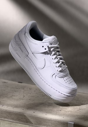 Baskets blanches Nike Air Force 1 avec lacets, bout perforé et logo sur le côté et la semelle, flottant au-dessus d'une surface en pierre texturée.