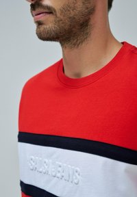 Camiseta de algodón roja con una franja horizontal blanca con el texto en relieve "SALSA JEANS" y acentos en azul marino, diseño de cuello redondo.