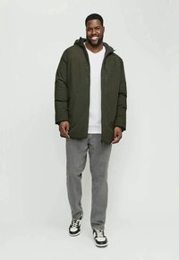 Homem sorridente a caminhar para a frente, vestindo uma jaqueta com capuz verde-escuro, camisa branca, calças cinzas e ténis preto e branco, contra um fundo simples.