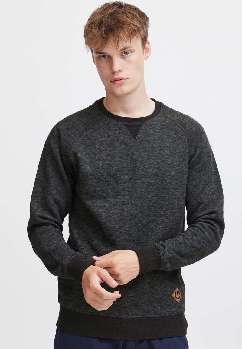 Czarny melanżowy sweatshirt z raglanowymi rękawami, okrągłym dekoltem, ściągaczami przy mankietach i u dołu. Posiada małą brązową naszywkę z logo w okolicy pasa.