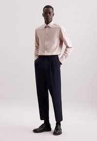 Jasnoróżowa koszula na guziki z długimi rękawami, w połączeniu z ciemnoniebieskimi plisowanymi spodniami i czarnymi eleganckimi butami. Prosty, nowoczesny design.