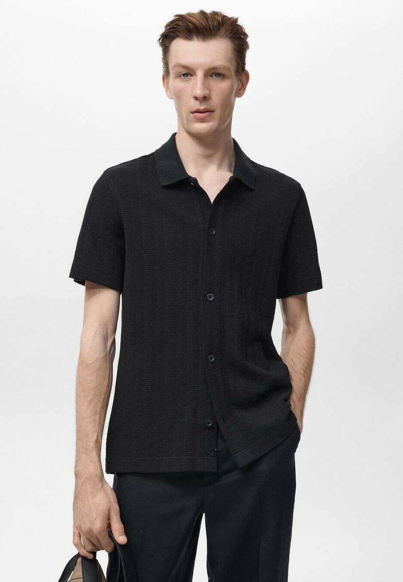 Zwart shirt met korte mouwen en een gestructureerd patroon, voorzien van een donkere kraag en knopenrij, gedragen met donkere broeken.