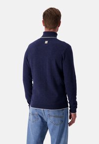 Maglione blu navy con colletto alto, polsini a costine e una banda bianca lungo l'orlo. Presenta un piccolo etichetta sulla zona sul retro del collo.
