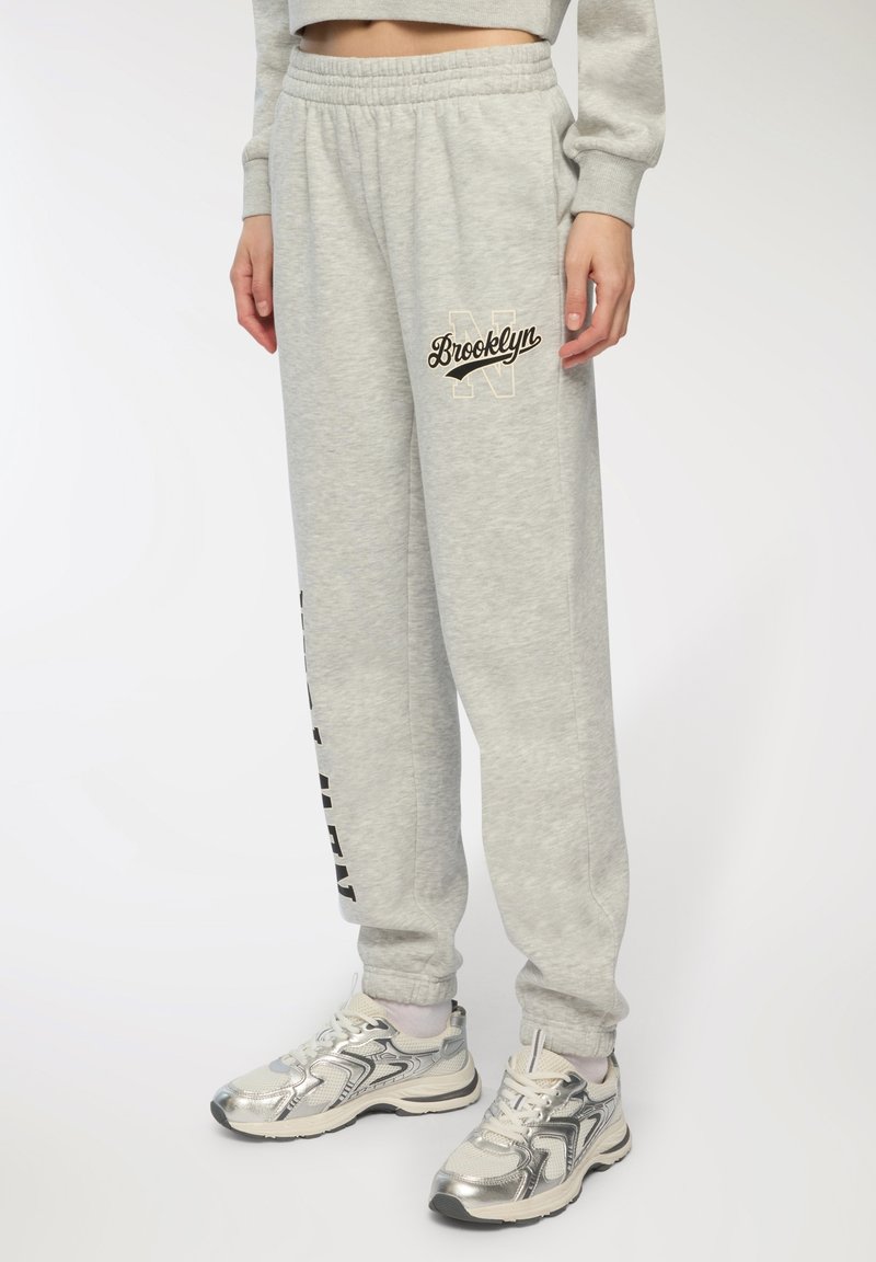Szare spodnie dresowe z elastycznym paskiem w talii i tekstem "Brooklyn" w kolorze czarnym, o luźnym kroju z czarnymi paskami po bokach.