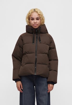 Junge Person mit kurzen lockigen blonden Haaren, die eine dunkelbraune gesteppte Jacke mit Kapuze und blaue Jeans vor hellem Hintergrund trägt.