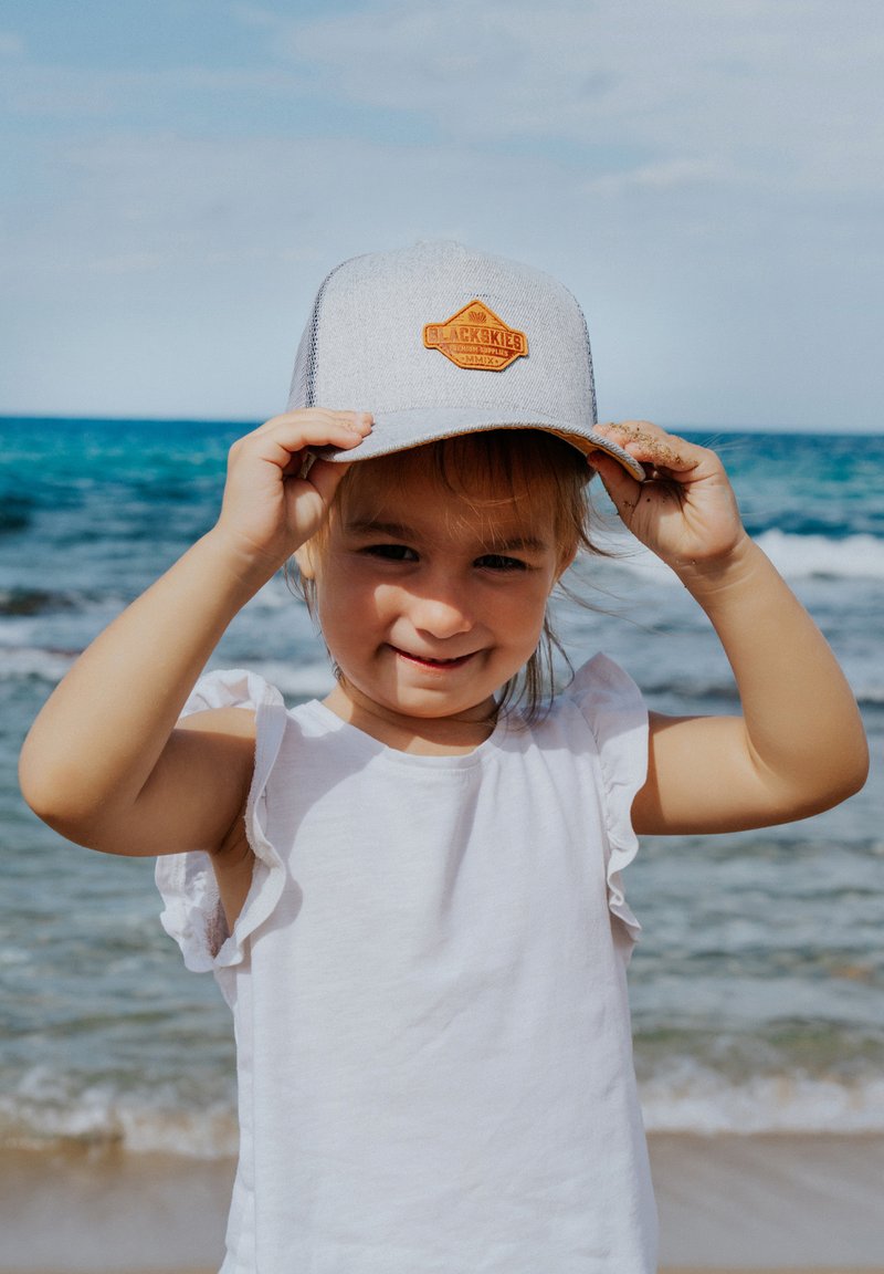 Graue Baseballmütze mit orangefarbenem Aufnäher, auf dem ein Logo zu sehen ist. Kind trägt ein weißes ärmelloses Shirt und steht am Strand mit dem Ozean im Hintergrund.