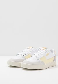 Białe trampki z żółtymi i szarymi akcentami, teksturowana gumowa podeszwa, materiały skórzane i zamszowe, perforowany nos, oraz detal z logo PUMA.