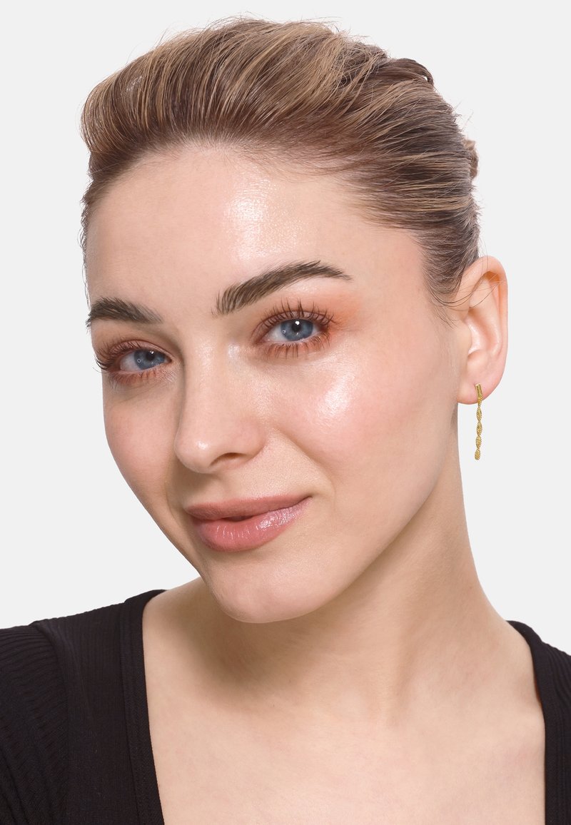 Goldene Creolen mit einem strukturierten Linkdesign. Das Model hat braune Haare, eine klare Haut und trägt ein schwarzes Oberteil vor einem schlichten Hintergrund.