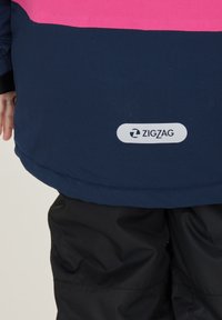 Marinejakke med pink accent, med et logo-tag der siger "ZIGZAG" på bagsiden. Stoffet ser glat ud med en let mat tekstur.