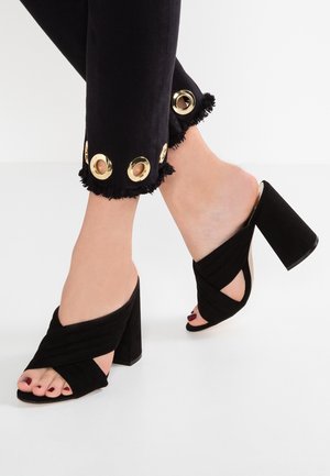 Sorte åbne mulepumps med krydsende design, lavet af blød ruskind, med en tyk blokhæl og en glat finish. Jeans har guldfarvede øjer.