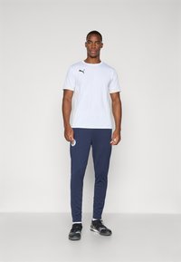 T-shirt de sport blanc avec logo noir, associé à un jogger bleu marine avec un patch circulaire. Modèle portant des chaussures de sport noires.