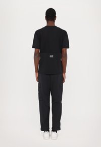 Black short-sleeve t-shirt with a solid back, featuring a logo at the waist, paired with black cargo pants and white sneakers.